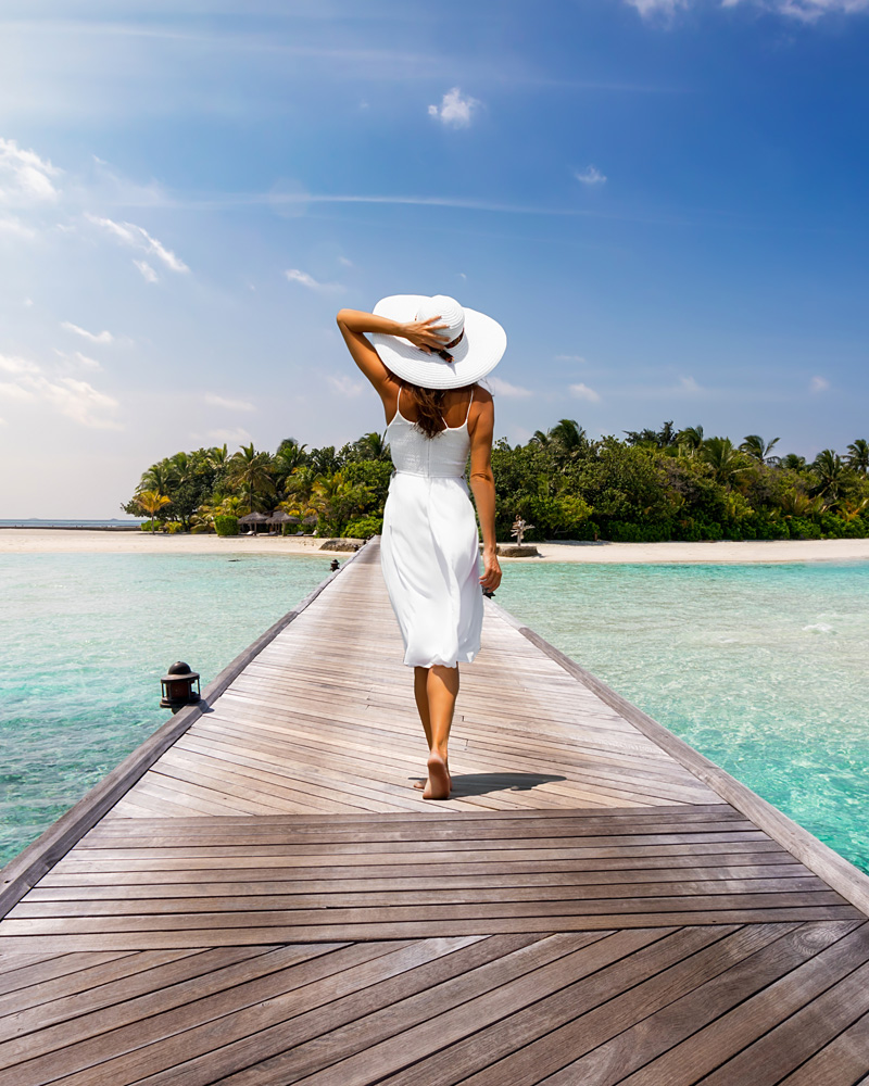 A woman walks away from the camera along a long wooden pier. She's dressed in a flowing white midi dress and wears a large wide-brimmed white sun hat, which she holds with one hand. She's barefoot on the weathered wooden planks.